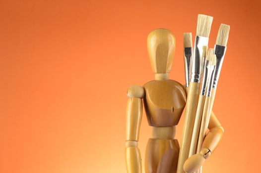 An Artists wooden Dummy Mannequin stands with some dry paintbrushes over an orange background.