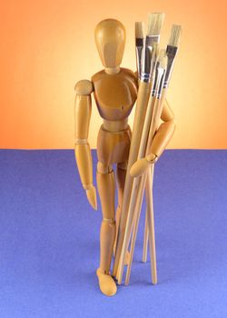 An Artists wooden Dummy Mannequin shot looking downward while holding some various dry paintbrushes over a blue and orange background.