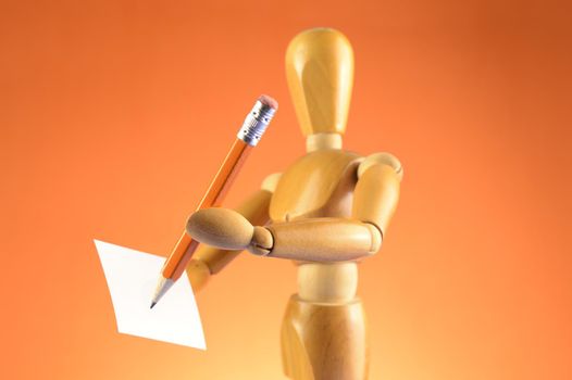 An Artists wooden Dummy Mannequin using a pencil and piece of paper to draw or write something of interest while over an orange color background.