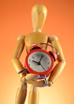 An Artists wooden Dummy Mannequin holds a red alarm clock over an orange background.