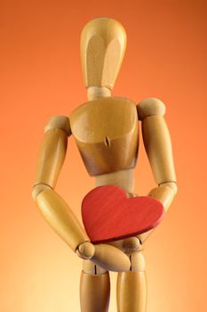 An Artist wooden Dummy Mannequin holds a red heart closely while standing over an orange background.