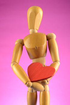 An Artist wooden Dummy Mannequin holds a red heart closely while standing over a pink background.