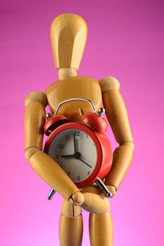An Artists wooden Dummy Mannequin holds a red alarm clock while standing over a pink background.