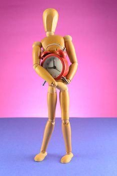 An Artist wooden Dummy Mannequin holds a red alarm clock closely while standing over a pink background.