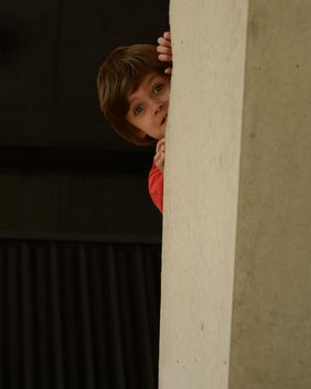 A young boy playfully plays peek a boo around a large cement pillar.