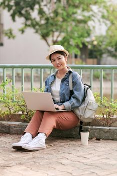 happy young traveler woman with backpack sitting in the city typing on her laptop, concept of technology, youth and digital nomad lifestyle, copy space for text