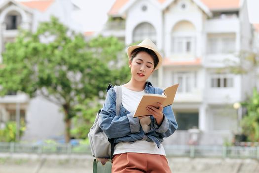 Asian female in casual clothes and beret with backpack standing on street reading book