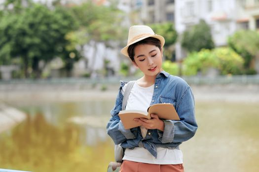 Asian female in casual clothes and beret with backpack standing on street reading book