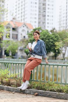 Pleased woman in casual wear holding in hand paper cup with hot drink while standing in city street at daytime