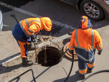 Workers over the open sewer hatch on a street. Repair of sewage, underground utilities, water supply system, water pipe accident