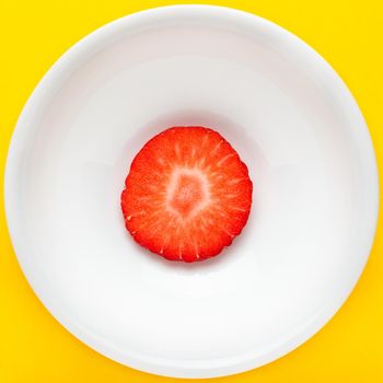 Slice of fresh strawberry on a round white plate on yellow background. Square image seen from above.