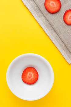 Three slices of fresh strawberries in a round white plate on yellow background and sackcloth. Vertical image viewed from above.