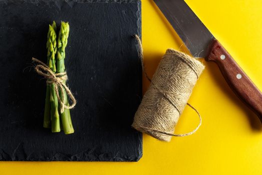 Asparagus. Bunch of fresh asparagus tied on a plate of slate with a knife and a roll of string on yellow background. Healthy vegetarian food. Horizontal image.