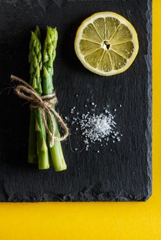 Asparagus. Bunch of fresh green asparagus with coarse salt and a slice of lemon on a slate plate on yellow background. Healthy vegetarian food. Vertical image