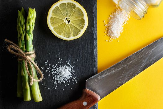 Asparagus. Bunch of fresh green asparagus with coarse salt, a slice of lemon and a knife on a plate of slate on yellow background. Healthy vegetarian food. Horizontal image