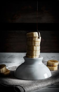Delicious dessert of fresh banana slices standing with liquid chocolate on top on a white bowl with slices around on a sackcloth and a background of wooden boards. Vertical image of dark moody style.