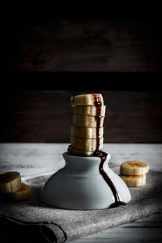 Delicious dessert of fresh banana slices standing with liquid chocolate on top on a white bowl with slices around on a sackcloth and a background of wooden boards. Vertical image of dark moody style.