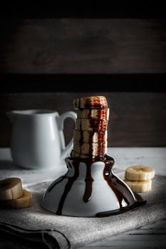 Delicious dessert of fresh banana slices standing with chocolate on top on a white bowl with slices around on a sackcloth and a background of wooden boards. Vertical image of dark moody style.