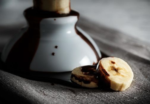 Delicious dessert of fresh banana slices with chocolate on top on a sackcloth seen from close up. Horizontal image of dark moody style.