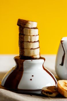 Delicious dessert of fresh banana slices standing with chocolate on top on a white bowl with slices around on a sackcloth and a yellow background. Vertical image.