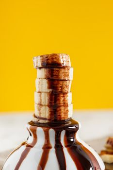 Delicious dessert of fresh banana slices standing with chocolate on top on a white bowl with slices around on a sackcloth and a yellow background. Vertical image.