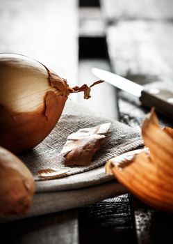 Still life of onion on a sackcloth with onion skins around and a knife in the background on wooden boards illuminated against the light. Dark moody style. Vertical image.