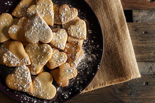 Delicious home-made heart-shaped cookies sprinkled with icing sugar on sackcloth and wooden boards. Horizontal image seen from above.