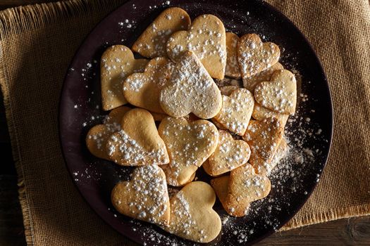 Delicious home-made heart-shaped cookies sprinkled with icing sugar on sackcloth and wooden boards. Horizontal image seen from above.