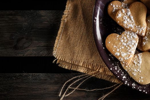 Delicious home-made heart-shaped cookies sprinkled with icing sugar on sackcloth and wooden boards. Horizontal image seen from above.