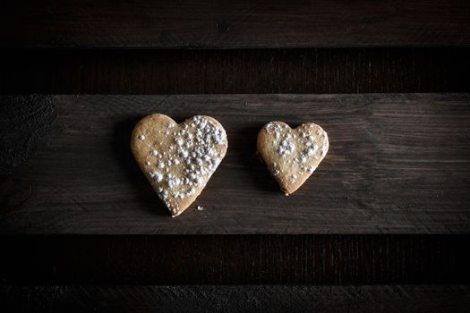 Two delicious home-made heart-shaped cookies sprinkled with icing sugar in a wooden board. Horizontal image seen from above. Concept of love in couple. Dark moody style.