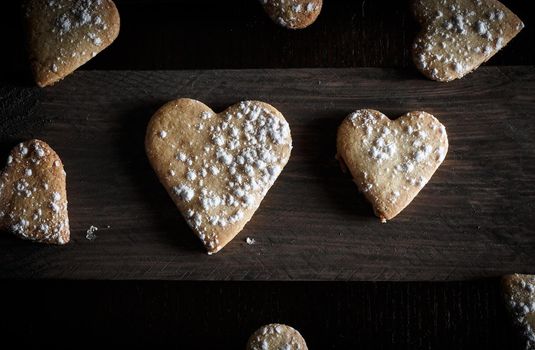 Delicious home-made heart-shaped cookies sprinkled with icing sugar in a wooden board. Horizontal image seen from above. Dark moody style.
