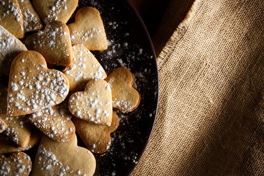 Delicious home-made heart-shaped cookies sprinkled with icing sugar on sackcloth and wooden boards. Horizontal image seen from above.