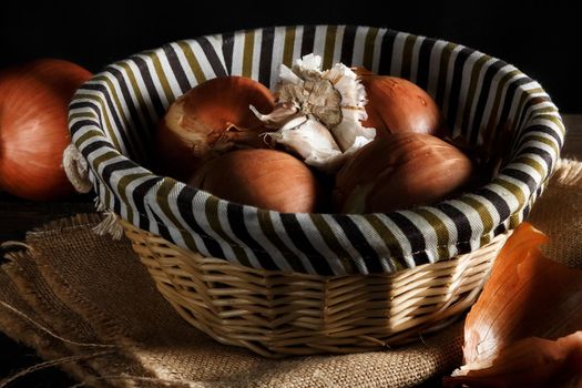 Onions and head of garlic in a wicker basket on wooden boards and sackcloth on black background. Rustic style. Horizontal image.