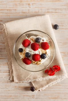 Raspberries, blueberries, cereals and yogurt in a glass bowl on sackcloth and wooden slats. Healthy breakfast for a healthy life. Vertical image view from above.