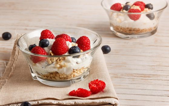 Raspberries, blueberries, cereals and yogurt in a glass bowl on sackcloth and wooden slats. Healthy breakfast for a healthy life. Horizontal image.