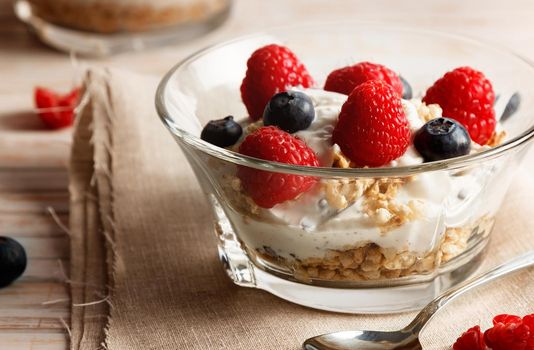 Raspberries, blueberries, cereals and yogurt in a glass bowl on sackcloth and wooden slats. Healthy breakfast for a healthy life. Horizontal image.