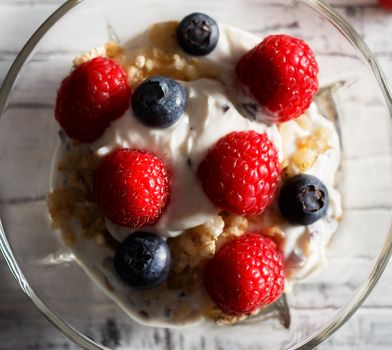 Raspberries, blueberries, cereals and yogurt in a glass bowl on wooden slats. Healthy breakfast for a healthy life. Square image view from above. Close up.