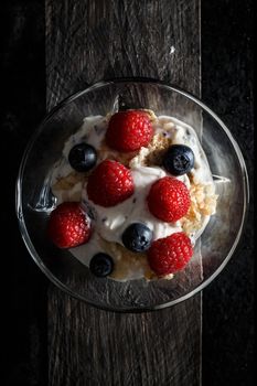 Raspberries, blueberries, cereals and yogurt in a glass bowl on old wooden boards. Healthy breakfast for a healthy life. Vertical image view from above.