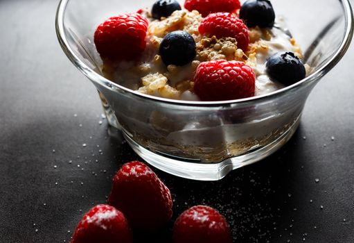 Raspberries, blueberries, cereals and yogurt in a glass bowl on a black surface. Healthy breakfast for a healthy life. Horizontal image.