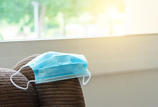 Surgical medical mask on top of a sofa and in front of a window with the sun behind.