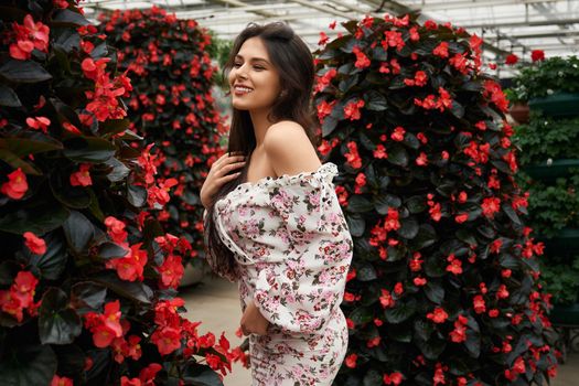 Smiling caucasian woman with long wavy hair posing near beautiful red flowers at greenhouse. Happy female model wearing romantic summer dress.