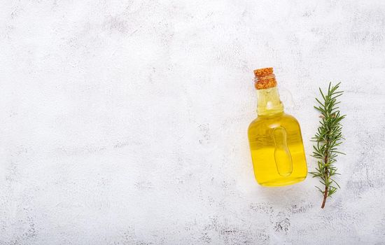 Glass bottle of olive oil and rosemary branch set up on white concrete background.