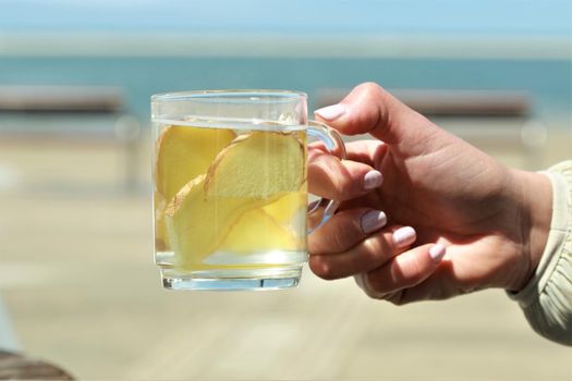 A glas of ginger tea with slices of ginger hold by a hand in front of a beach