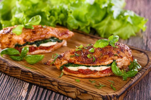 Baked chicken breast with mozzarella, sun-dried tomatoes, spinach. The meat is served on a wooden board with basil and lettuce.