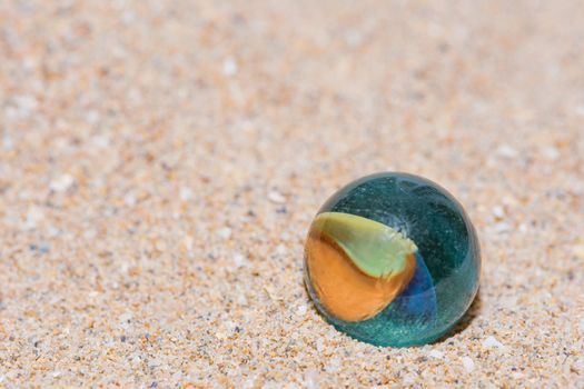 Glass marble with colored interior resting on the sand, close-up image of a playful object