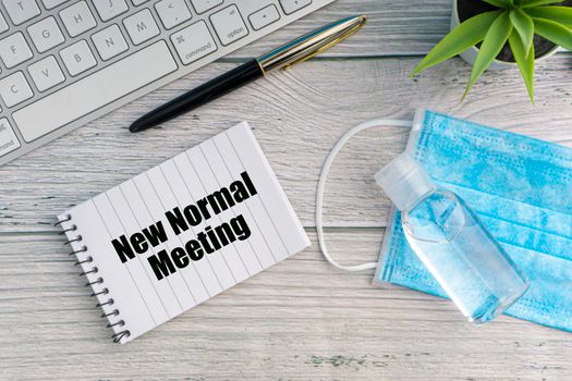 NEW NORMAL MEETING text with notebook, safety face mask, keyboard, fountain pen, hand sanitizer and decorative plant on wooden background. Covid 19 and coronavirus concept