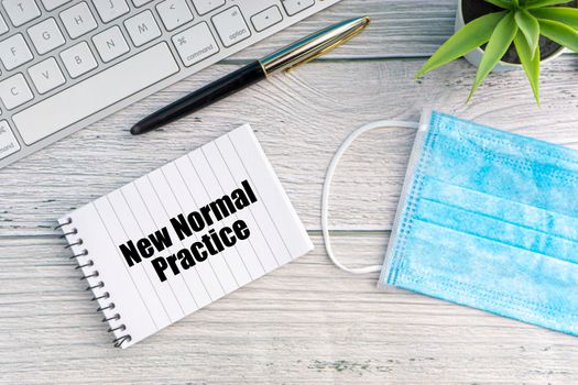 NEW NORMAL PRACTICE text with notebook, safety face mask, keyboard, fountain pen and decorative plant on wooden background. Covid 19 and coronavirus concept
