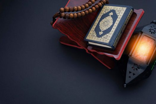 Holy Al Quran with written arabic calligraphy meaning of Al Quran, lantern lamp and rosary beads or tasbih on black background.