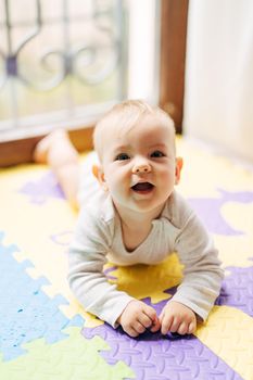Surprised joyful baby lies on his tummy on a colored rug on the floor against the background of a window. High quality photo