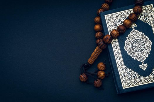 Holy Al Quran with written arabic calligraphy meaning of Al Quran and rosary beads or tasbih on black background. Copy space and crop fragment